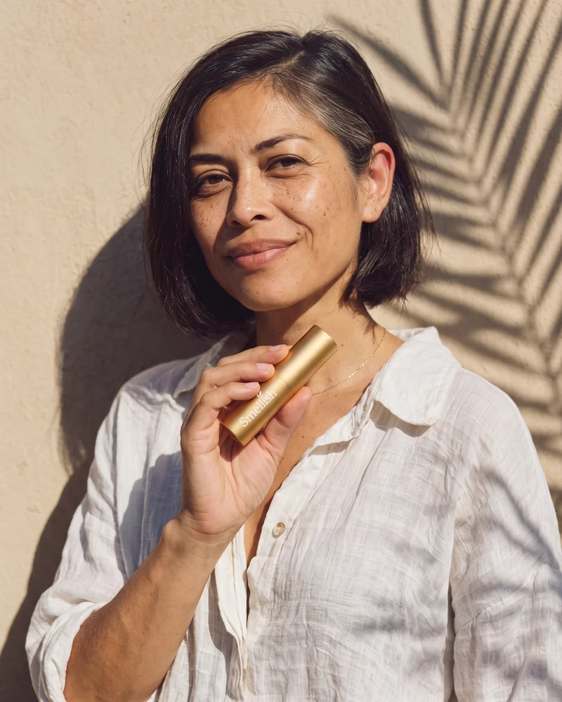 Vrouw in een wit linnen blouse houdt een gouden Smellish twist-up vast tegen een zonnige muur met palmschaduw