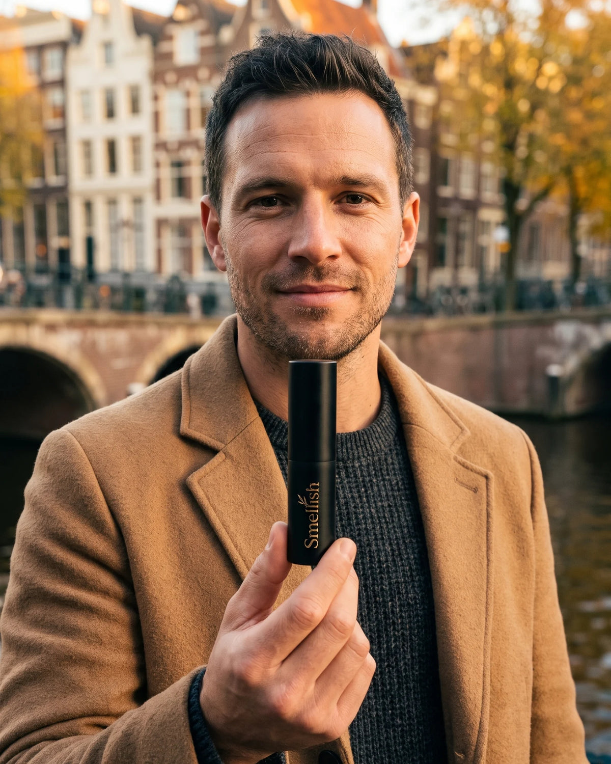 Man in camelkleurige jas houdt een zwarte Smellish twist-up naar de camera bij een Amsterdamse gracht in de herfst