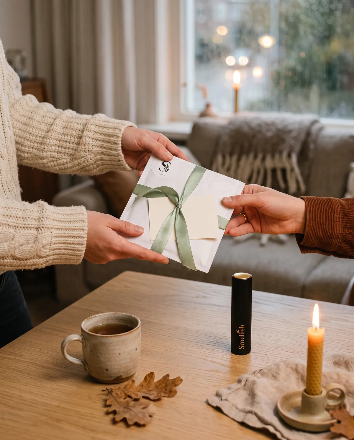 Handen geven een witte cadeau-envelop met groen lint door, met een zwarte twist-up travelspray op de salontafel